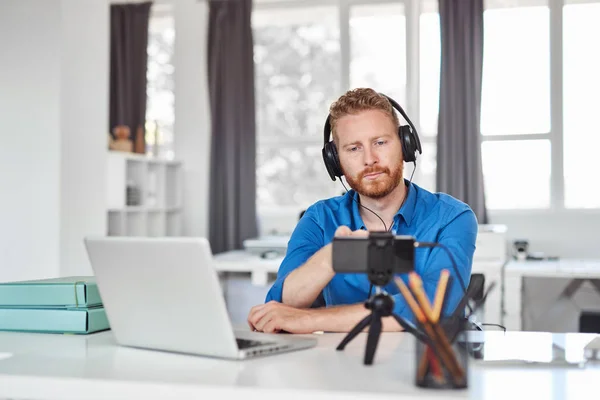 Joven empleado caucásico que tiene videollamada por teléfono inteligente mientras está sentado en la oficina. En la cabeza hay auriculares y en el escritorio es portátil y café . — Foto de Stock