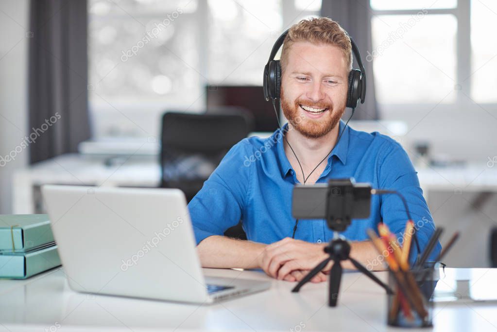 Young Caucasian employee having video call over smart phone while sitting in office. On head are headphones and on desk is laptop and coffee.