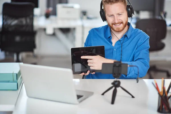 Joven empleado trabajador caucásico que tiene videollamada por teléfono inteligente. En las manos es tableta y en la cabeza son auriculares. Concepto de negocio remoto . — Foto de Stock