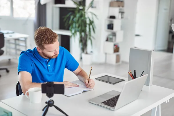 Empleado caucásico dedicado a trabajar duro sentado en una oficina moderna y escribiendo algo en un cuaderno . — Foto de Stock