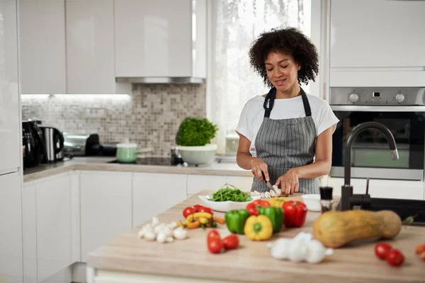 Linda dona de casa de raça mista em avental de pé na cozinha e cortando cogumelos. No balcão da cozinha estão todos os tipos de legumes . — Fotografia de Stock