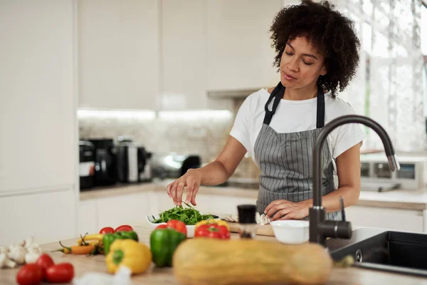Attraktiv blandad ras kvinna i förkläde stående i köket och med raket från plattan. På köksbänken finns tomater, paprika och zucchini. — Stockfoto