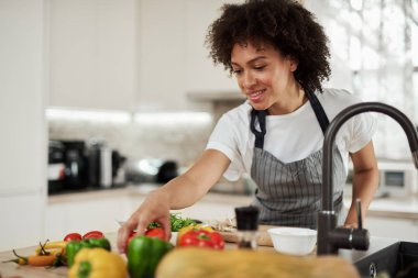 Mutfakta dururken domates için ulaşan önlük güzel karışık ırk ev kadını. Mutfak tezgahında sebzeler var. Akşam yemeği hazırlığı.