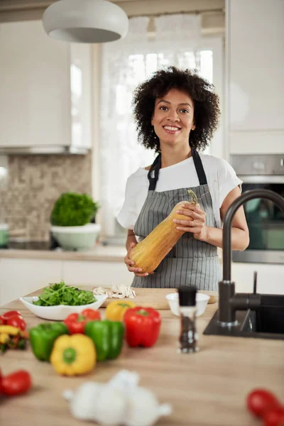Vacker blandad ras hemmafru i förkläde stående i köket och hålla zucchini på köksbänken är grönsaker. Måltids beredning. — Stockfoto