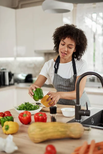 Mooie gemengde race huisvrouw in schort bereiken voor peper terwijl staande in de keuken. Op de keuken teller zijn groenten. Voorbereiding van het diner. — Stockfoto