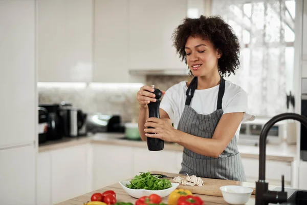 Aantrekkelijke gemengde race vrouw in grijze schort het toevoegen van peper in schaal met rucola en champignons. Op de keuken teller zijn verschillende groente. — Stockfoto