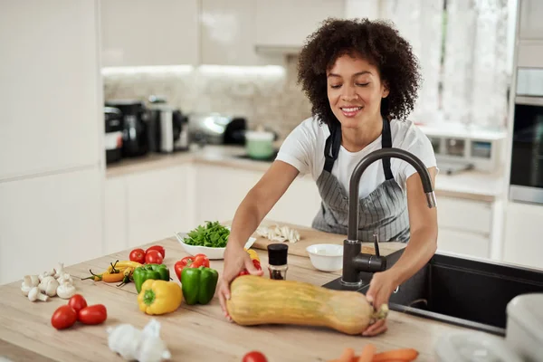 Attractive mixed race woman in apron taking zucchini from kitchen counter. On kitchen counter are lots of vegetables.