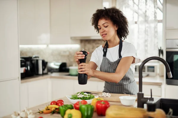 Приваблива змішана жінка в сірому фартусі додає перець у миску з ракетами та грибами. На кухонній стільниці різні овочі . — стокове фото