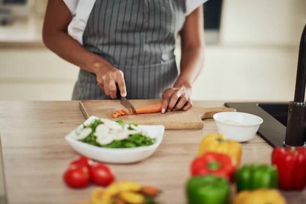 Gros plan de la femme métissée dans le tablier de coupe de carotte pour le repas. Sur le comptoir de cuisine sont des légumes . — Photo