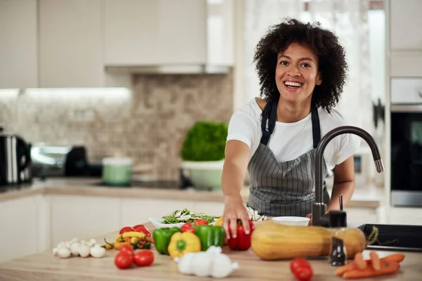 Beautiful mixed race housewife reaching for red pepper while standing in kitchen. Dinner preparation.