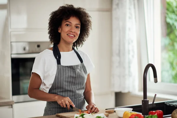 Attraktiv blandras kvinna i förkläde skära morot och laga middag när du står i köket. På köksbänken finns grönsaker. — Stockfoto