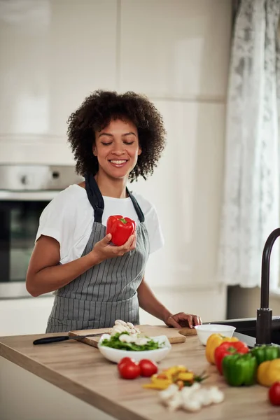 Приваблива змішана гоночна домогосподарка в фартусі, що тримає червоний перець, стоячи на кухні. На кухонній стільниці є овочі . — стокове фото