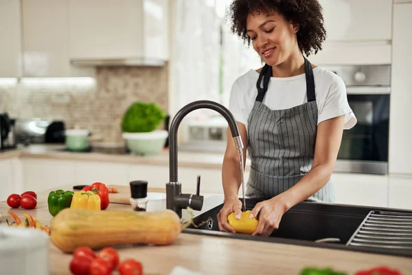 Charmant gemengd ras vrouw in schort staan in de keuken en wassen gele paprika in gootsteen. Op het aanrecht zijn verschillende soorten groenten. — Stockfoto