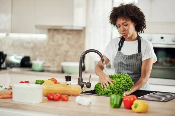 Mooie gemengde ras vrouw met krullend haar en in schort staan in de keuken en wassen salade in gootsteen. — Stockfoto