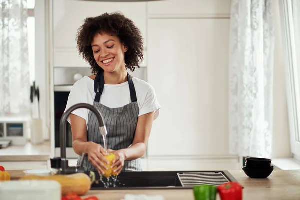 Charmig blandras kvinna i förkläde stående i kök annons tvätta gula paprika i handfat. På köksbänken finns olika sorters grönsaker. — Stockfoto