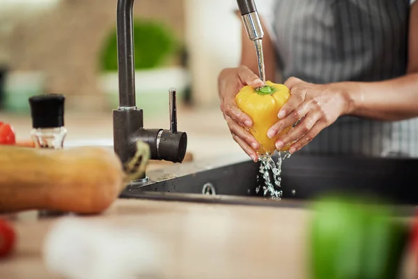 Önlüklü, mutfak reklamında durup lavaboda sarı biber yıkayan melez bir kadın.. — Stok fotoğraf