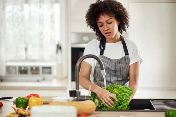 Mooie gemengde ras vrouw met krullend haar en in schort staan in de keuken en wassen salade in gootsteen. — Stockfoto