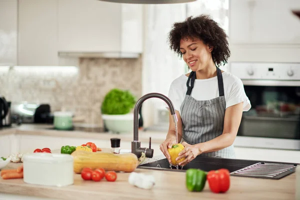 Чарівна змішана расова жінка в фартусі, що стоїть на кухні, миючи жовту паприку в раковині. На кухонній стільниці є різні види овочів . — стокове фото
