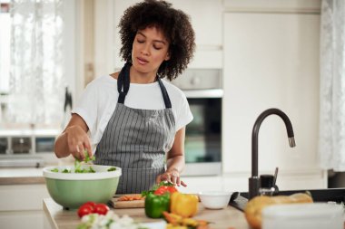 Önlüklü çekici melez kadın mutfakta duruyor ve kaseye doğranmış marul koyuyor. Yemek hazırlama konsepti.
