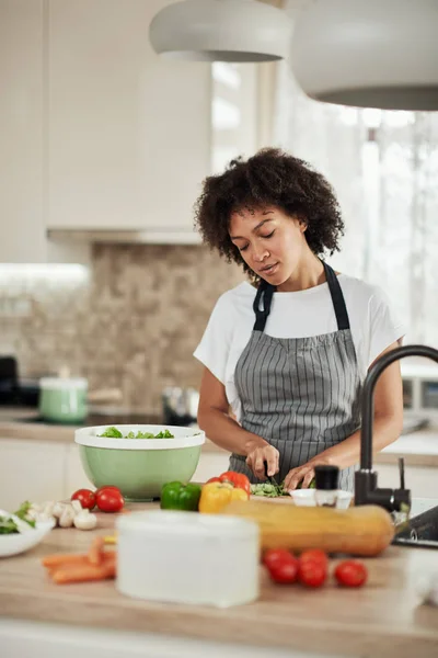 Attraktiv blandras kvinna i förkläde skära sallad för sallad. Köksinredning. — Stockfoto