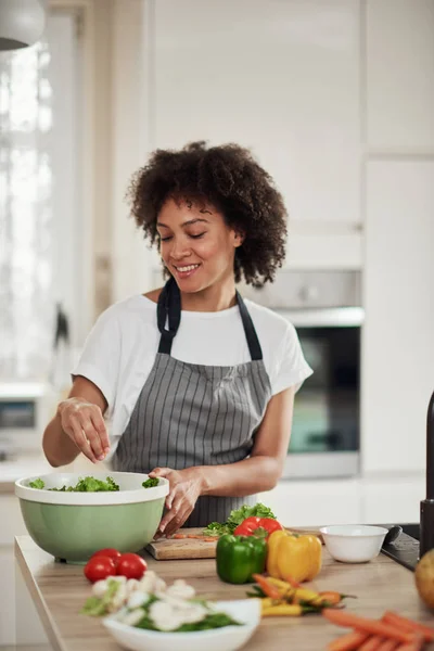 Mulher de raça mista atraente em avental em pé na cozinha e colocando alface picada na tigela. Conceito de preparação de refeições . — Fotografia de Stock
