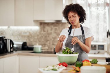 Çekici melez, kıvırcık saçlı, önlüklü, yerli mutfakta salata karıştıran bir kadın..