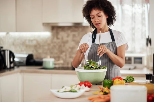 Mulher de raça mista atraente com cabelo encaracolado e em avental misturando salada enquanto em pé na cozinha doméstica . — Fotografia de Stock