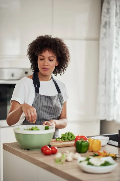 Mulher de raça mista atraente em avental em pé na cozinha e colocando alface picada na tigela. Conceito de preparação de refeições . — Fotografia de Stock