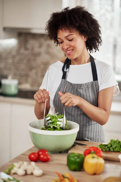 Mulher de raça mista atraente com cabelo encaracolado e em avental misturando salada enquanto em pé na cozinha doméstica . — Fotografia de Stock