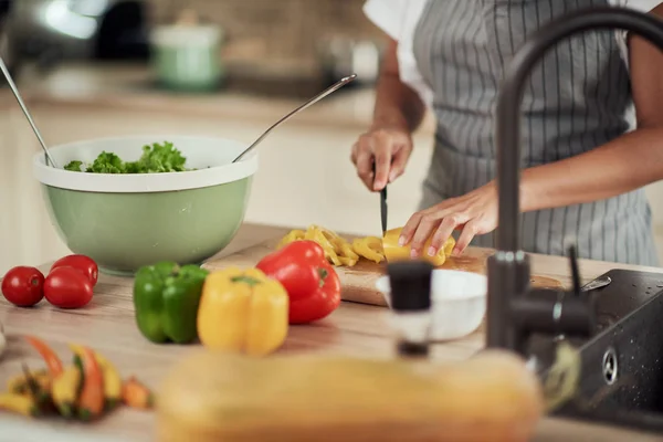 Close-up van gemengde ras vrouw in schort snijden gele peper voor het diner. Keuken interieur. Op het aanrecht staan pepers, courgettes en tomaten. — Stockfoto