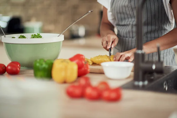 Gros plan d'une femme métisse en tablier coupant du poivron jaune pour le dîner. Intérieur de la cuisine. Sur le comptoir de la cuisine sont poivrons, courgettes et tomates . — Photo