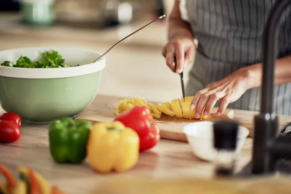 Primer plano de la mujer de raza mixta en delantal de corte de pimiento amarillo para la cena. Interior de la cocina. En el mostrador de la cocina son pimientos, calabacín y tomates . — Foto de Stock