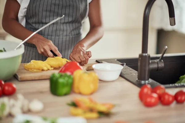 Close up de mulher de raça mista em avental de corte de pimenta amarela para o jantar. Interior da cozinha. No balcão da cozinha estão pimentas, abobrinha e tomates . — Fotografia de Stock