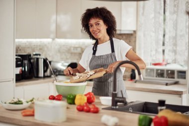Önlüklü, kıvırcık saçlı, evde mutfakta sebze karıştıran çekici bir ırk kadını..