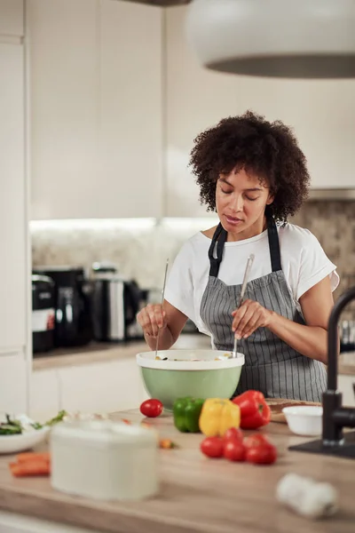 Mulher de raça mista atraente em avental e com cabelos cacheados misturando legumes em tigela enquanto estava na cozinha em casa . — Fotografia de Stock