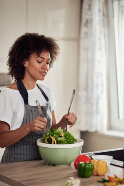 Önlüklü, kıvırcık saçlı, evde mutfakta sebze karıştıran çekici bir ırk kadını..