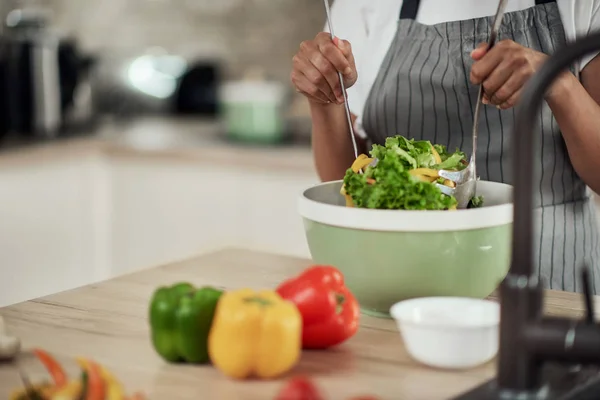 Aantrekkelijke gemengde ras vrouw in schort mengen van groenten in kom terwijl staan in de keuken thuis. — Stockfoto