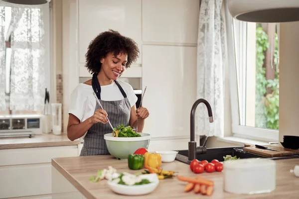 Приваблива змішана раса жінка в фартуху, змішуючи овочі в мисці, стоячи на кухні вдома . — стокове фото