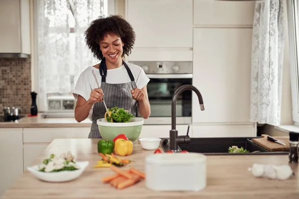 Приваблива змішана раса жінка в фартуху, змішуючи овочі в мисці, стоячи на кухні вдома . — стокове фото