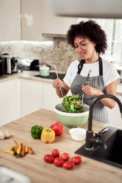 Attraktiv blandras kvinna i förkläde blanda grönsaker i skål när du står i köket hemma. — Stockfoto