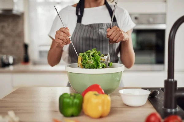 Aantrekkelijke gemengde ras vrouw in schort mengen van groenten in kom terwijl staan in de keuken thuis. — Stockfoto