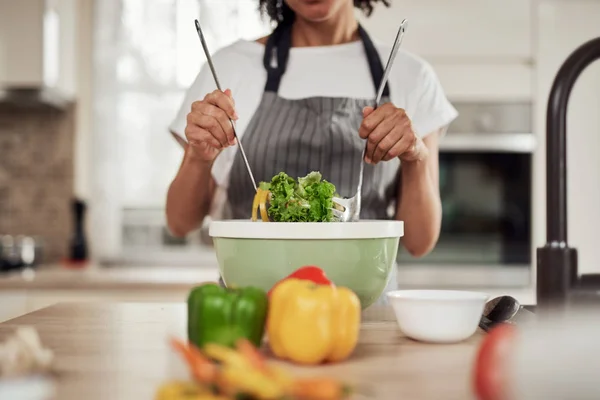 Aantrekkelijke gemengde ras vrouw in schort mengen van groenten in kom terwijl staan in de keuken thuis. — Stockfoto