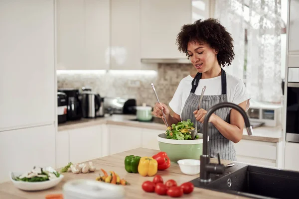 Attraktiv blandras kvinna i förkläde blanda grönsaker i skål när du står i köket hemma. — Stockfoto