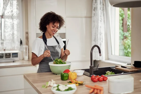 Mulher de raça mista atraente em avental misturando legumes na tigela enquanto estava na cozinha em casa . — Fotografia de Stock
