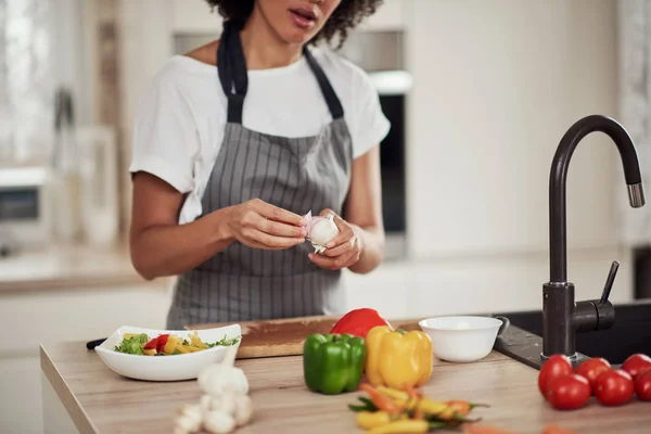 Prachtige gemengde ras vrouw in schort staan in de keuken en peeling knoflook. — Stockfoto