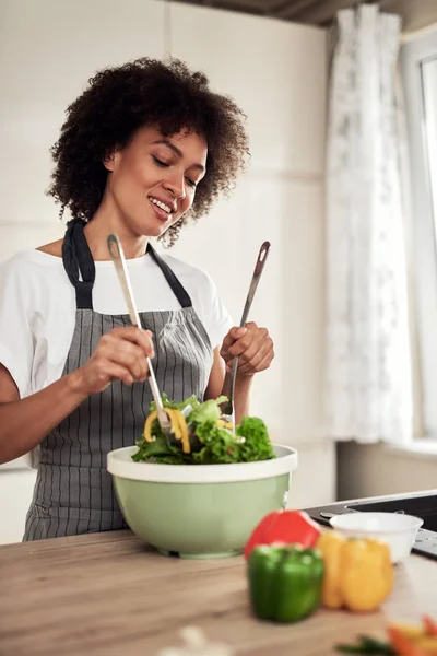 Aantrekkelijke gemengde ras vrouw in schort mengen van groenten in kom terwijl staan in de keuken thuis. — Stockfoto