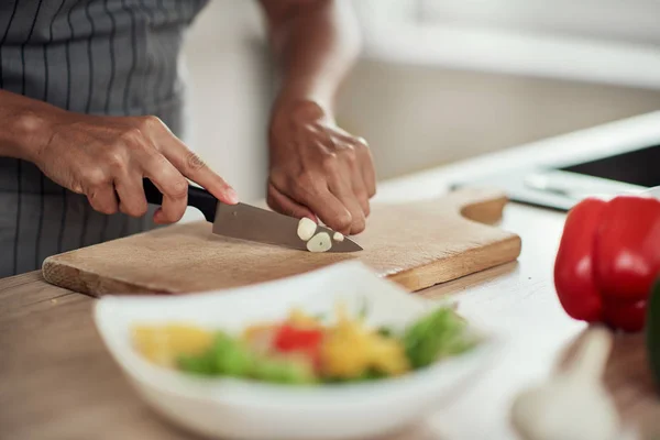 Primer plano de la mujer de raza mixta en delantal de pie en la cocina doméstica y cortar el ajo . — Foto de Stock