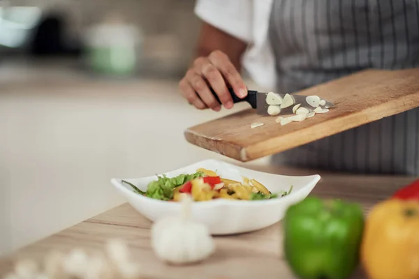 Cerrar mujer de raza mixta en delantal sosteniendo tabla de cortar y poner el ajo picado en un tazón con verduras . — Foto de Stock