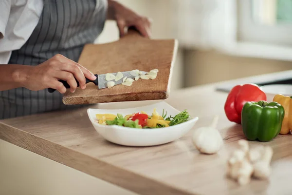Cerrar mujer de raza mixta en delantal sosteniendo tabla de cortar y poner el ajo picado en un tazón con verduras . — Foto de Stock