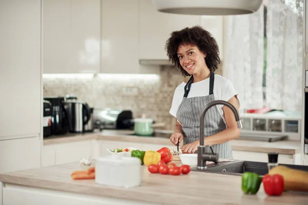 Gorgeous mixed race housewife in apron standing in kitchen and cGorgeous mixed race housewife in apron standing in kitchen and chopping mushrooms. On kitchen counter are all sorts of vegetables. — Stockfoto